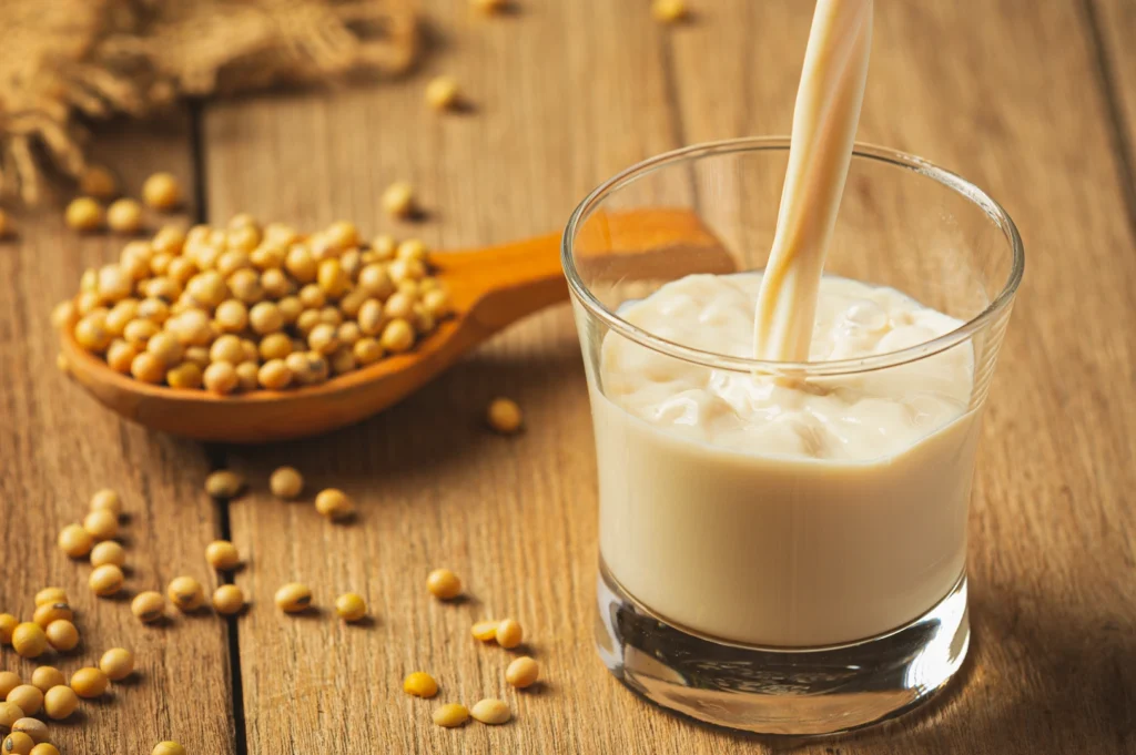 Close-up shot of creamy soy milk being poured into a clear glass on a rustic wooden table, with whole soybeans scattered beside a wooden spoon in natural light.