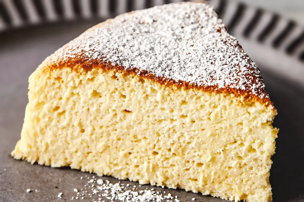 Light and airy Japanese‑style soufflé cheesecake slice dusted with powdered sugar on a dark serving plate