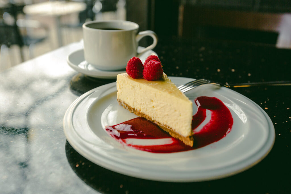 Slice of creamy cheesecake topped with fresh raspberries and served with a raspberry sauce swirl beside a cup of coffee in a café setting