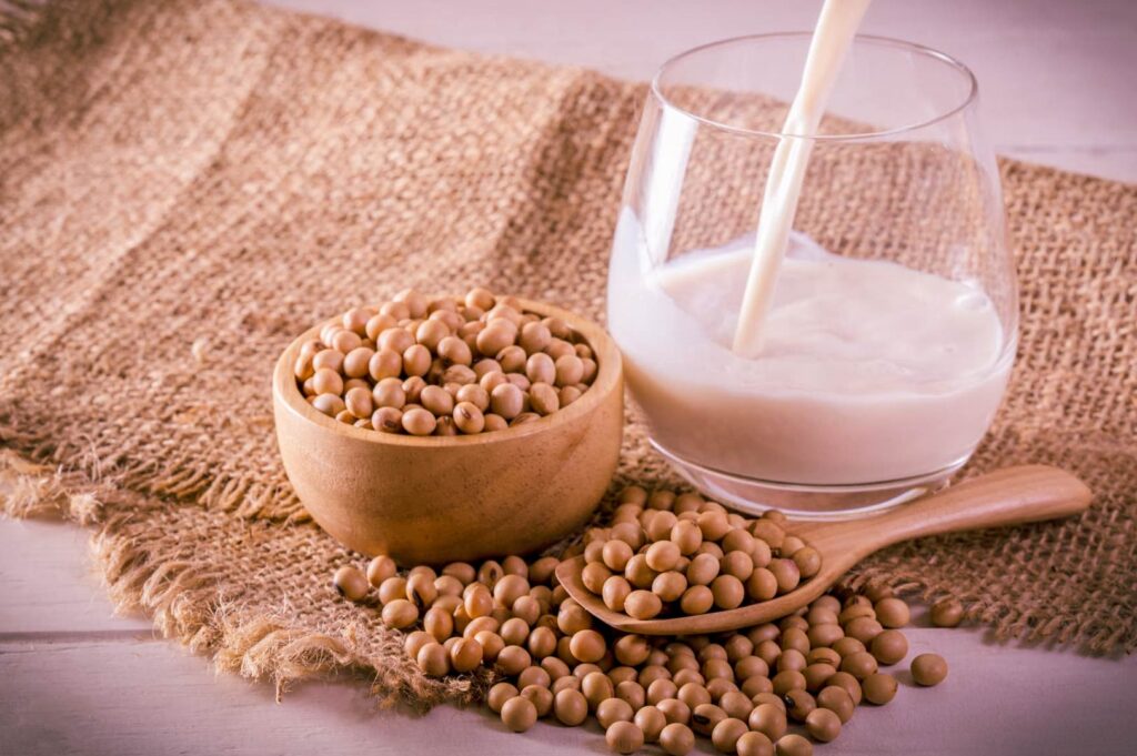 Bowl of dried soybeans beside a glass of freshly poured soy milk on a rustic burlap cloth.