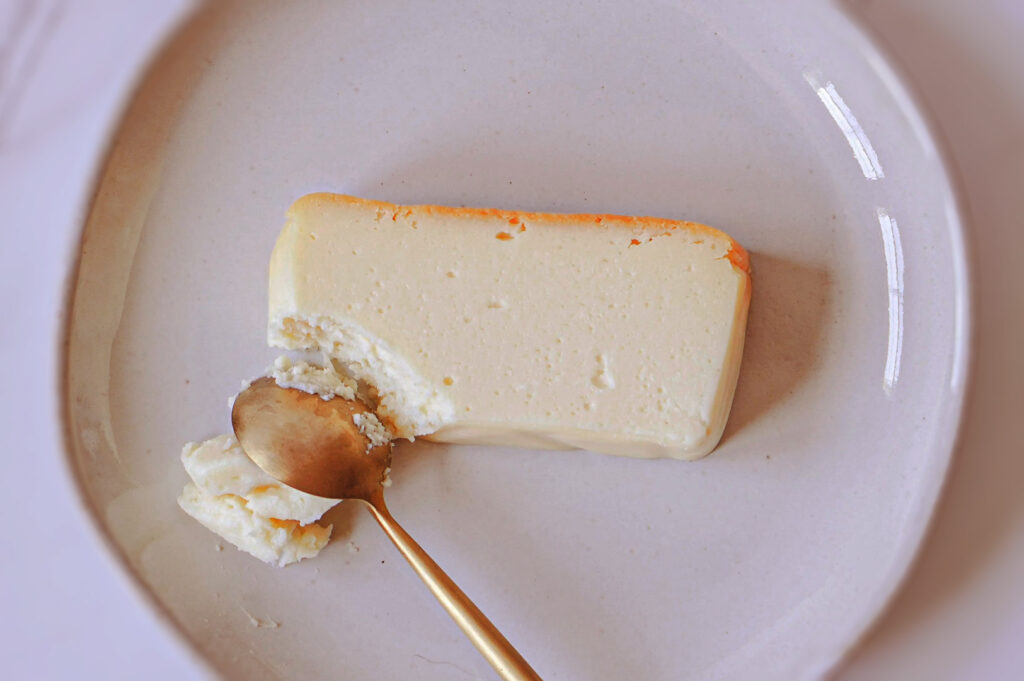 A piece of soft, creamy cheesecake on a plate with a spoon taking a portion from the slice.