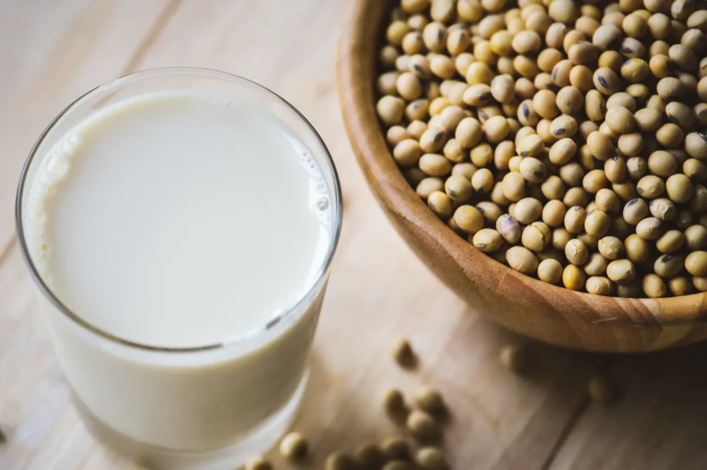 Overhead flat-lay shot of a glass of creamy soy milk beside a wooden bowl filled with whole soybeans on a natural wooden surface.