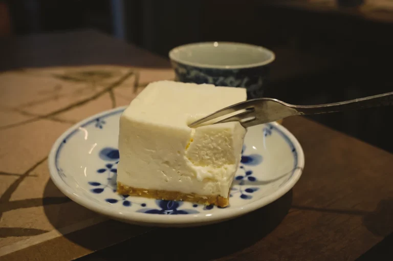 Close-up three-quarter angle shot of a fork cutting into a creamy soy cheesecake slice on a ceramic plate, revealing dense yet soft dairy-free texture made from soy.