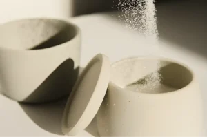Two minimalist ceramic cups, one with a lid, sit on a surface with salt being poured into the open cup, casting soft shadows in gentle lighting.