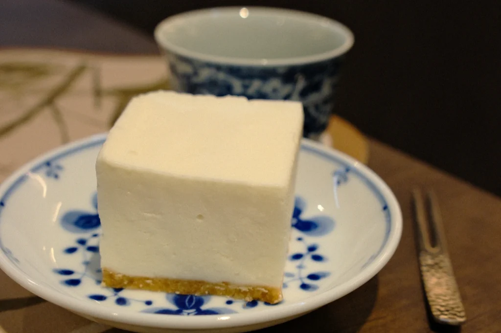 A slice of white cheesecake on a blue floral plate with a graham cracker crust, accompanied by a cup with a similar blue pattern on a wooden table.