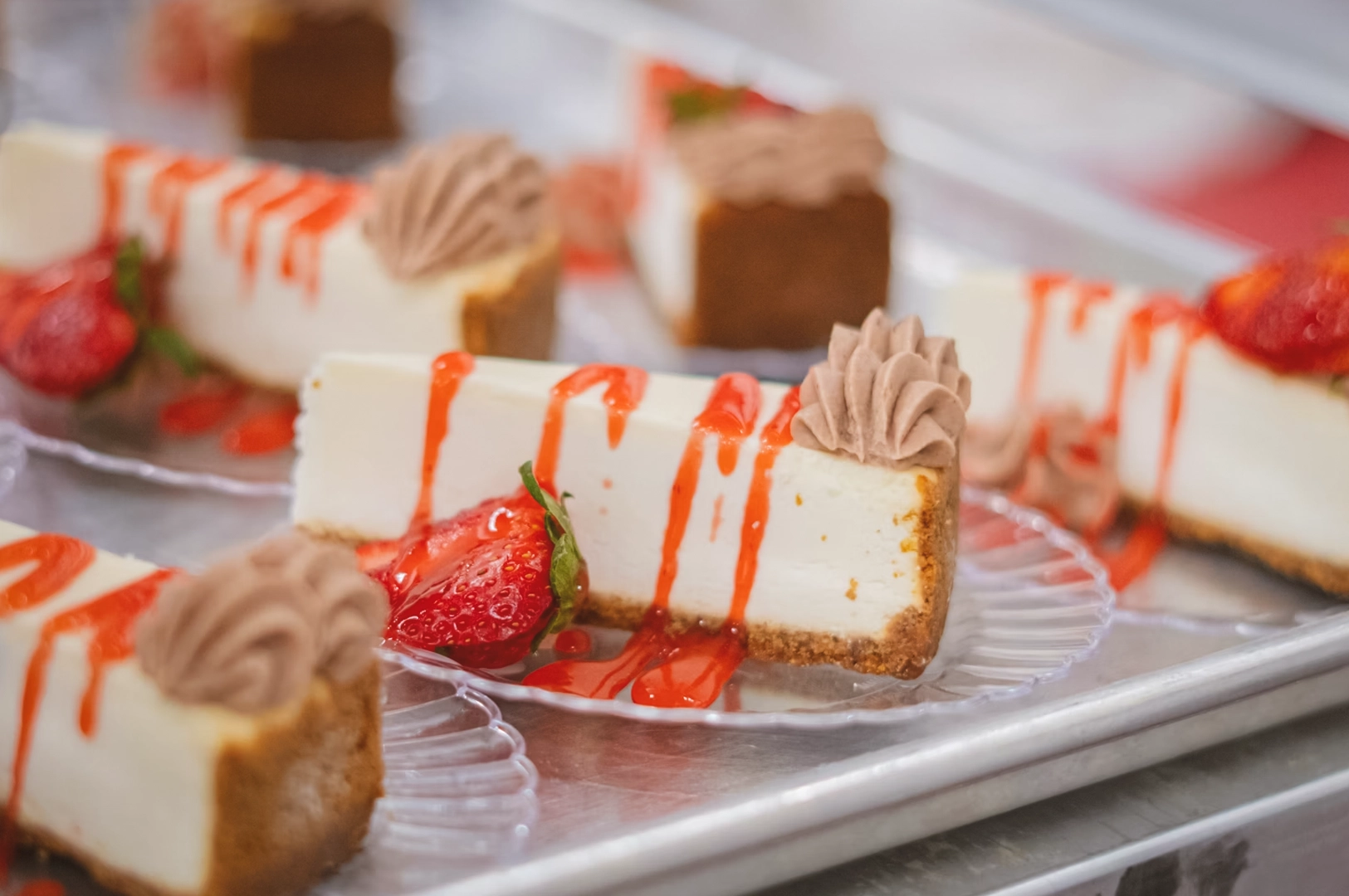 Eye-level close-up of sliced cheesecake topped with whipped cream, strawberries, and strawberry sauce, arranged on glass plates in a bakery display, illustrating rich dessert indulgence.