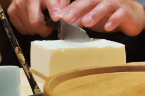 Close-up of hands carefully slicing a block of soft tofu with a knife on a wooden cutting board, conveying a sense of precision and focus.