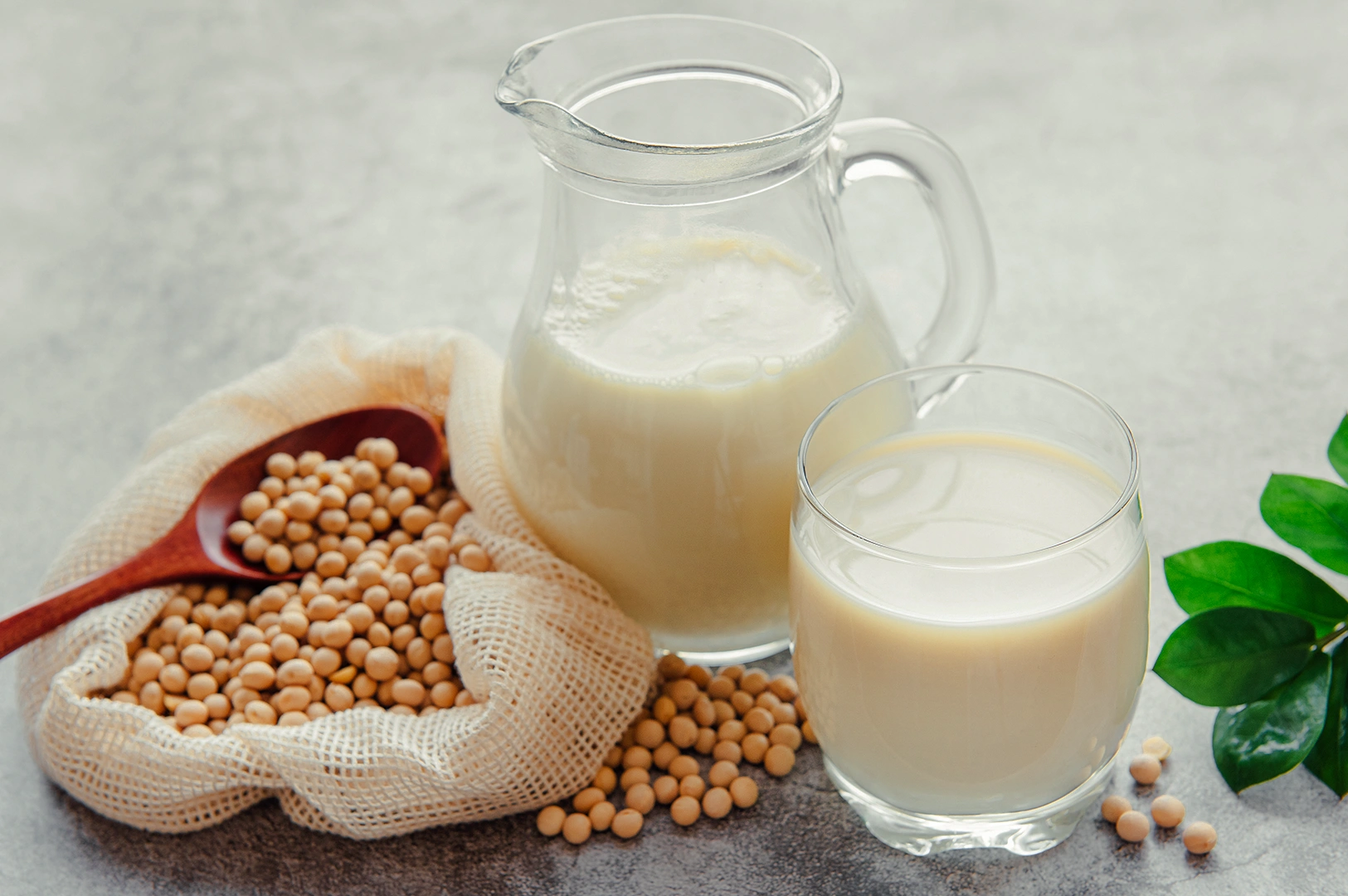 Eye‑level wide close‑up shot of soy milk in a glass and pitcher beside whole soybeans in a cloth sack, arranged on a neutral surface to showcase plant‑based dairy ingredients.