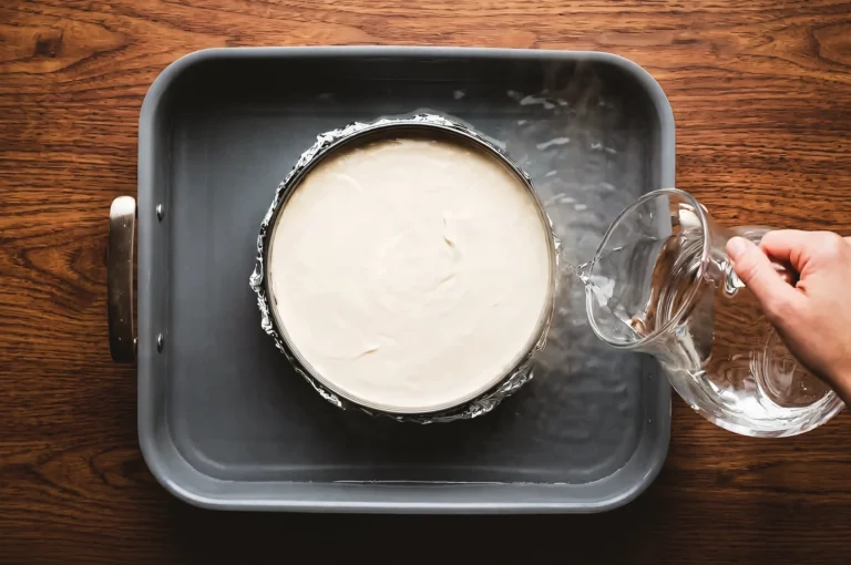 Top-down overhead shot of a cheesecake pan wrapped in foil and placed inside a larger roasting tray, with water being poured around it to prepare a water bath for even baking.