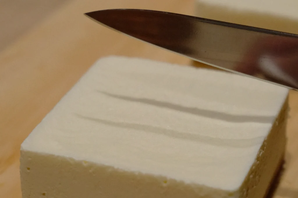 A close-up of a silver knife slicing through a smooth, white block of tofu on a wooden cutting board, highlighting a clean, precise cut.