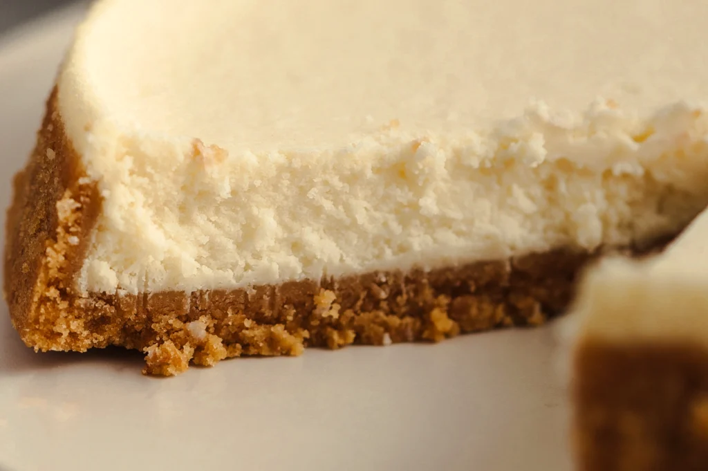 Extreme close-up side-angle shot of a creamy baked cheesecake slice with a smooth vanilla filling and crumbly biscuit base, highlighting rich texture and classic cheesecake layers.