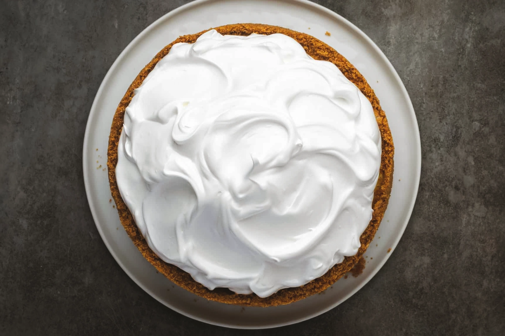 Top-down overhead shot of a whole cheesecake covered with a thick layer of whipped cream on a white plate, emphasizing richness and density in dessert presentation.