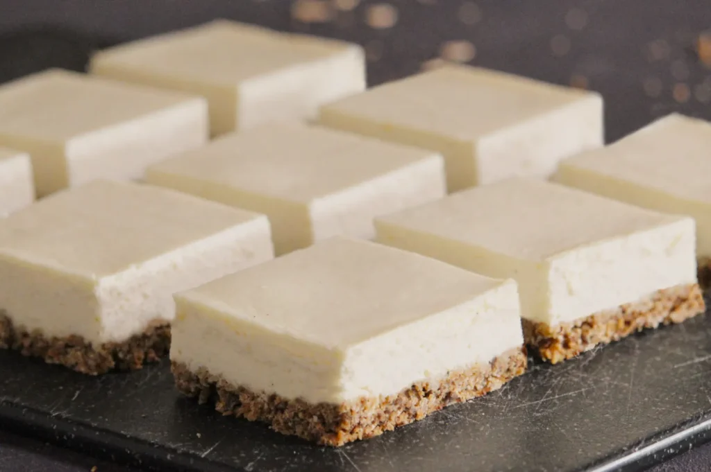 Nine neatly arranged square cheesecake slices with creamy tops and crumbly crusts are displayed on a dark slate, creating an inviting, delicious scene.
