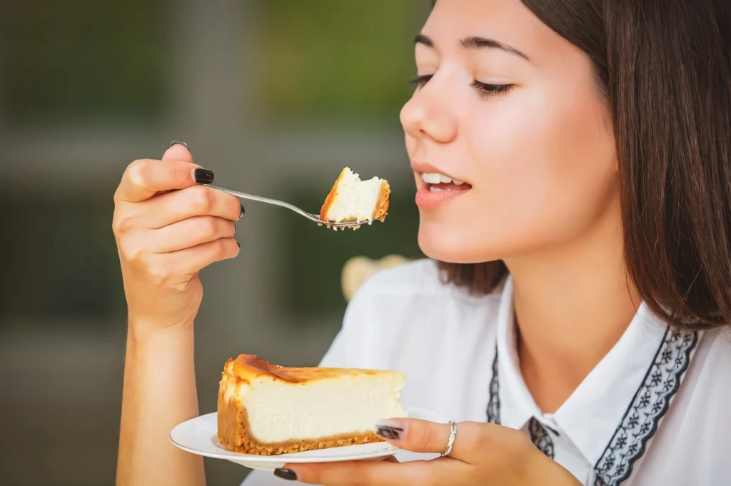 Three-quarter close-up café dining shot of a person holding a forkful of cheesecake while a cheesecake slice rests on a white plate, highlighting creamy texture, golden crust, and casual café dessert enjoyment in natural light.