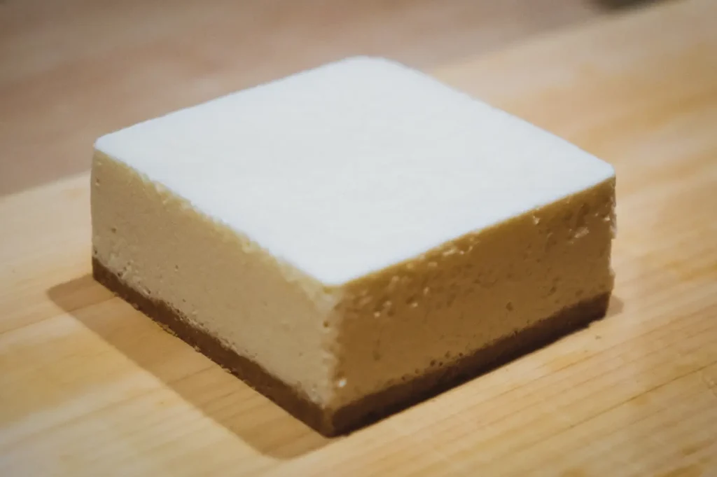 Close-up three-quarter angle shot of a square soybean cheesecake on a wooden cutting board, showing smooth white soy-based cheesecake filling over a light brown crust.