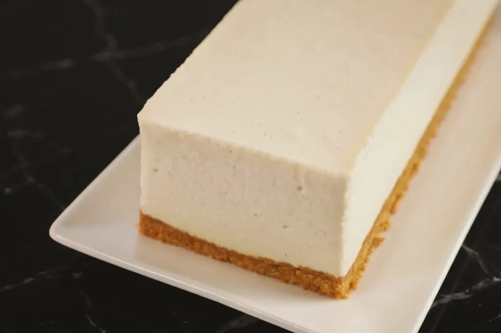 Eye-level side-angle shot of a rectangular soy cheesecake with a smooth white filling and a thin crumb crust, served on a white plate against a dark background.