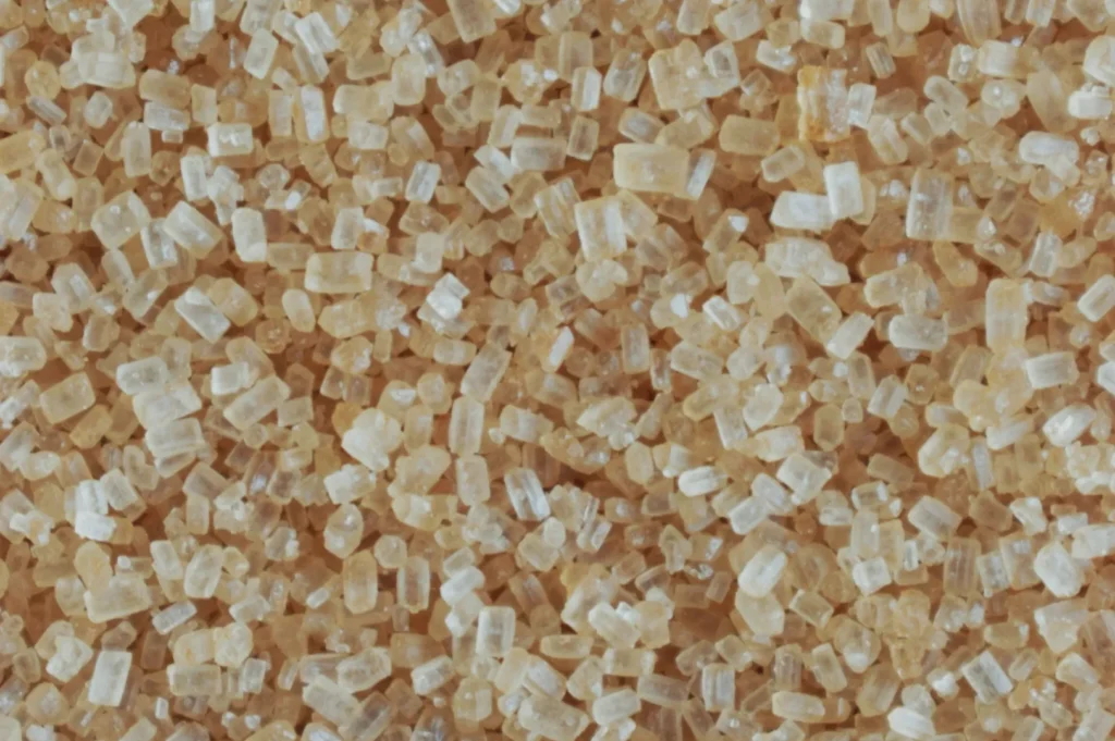 Close-up of granulated brown sugar crystals. The image shows a textured mass of irregular, amber-colored grains, creating a rustic and natural feel.