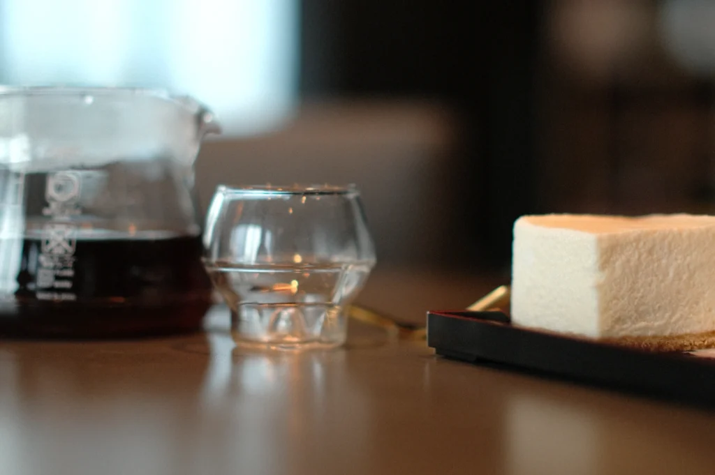 A clear glass coffee carafe, a small glass, and a slice of cake on a tray sit on a wooden table. The scene conveys a cozy, inviting atmosphere.
