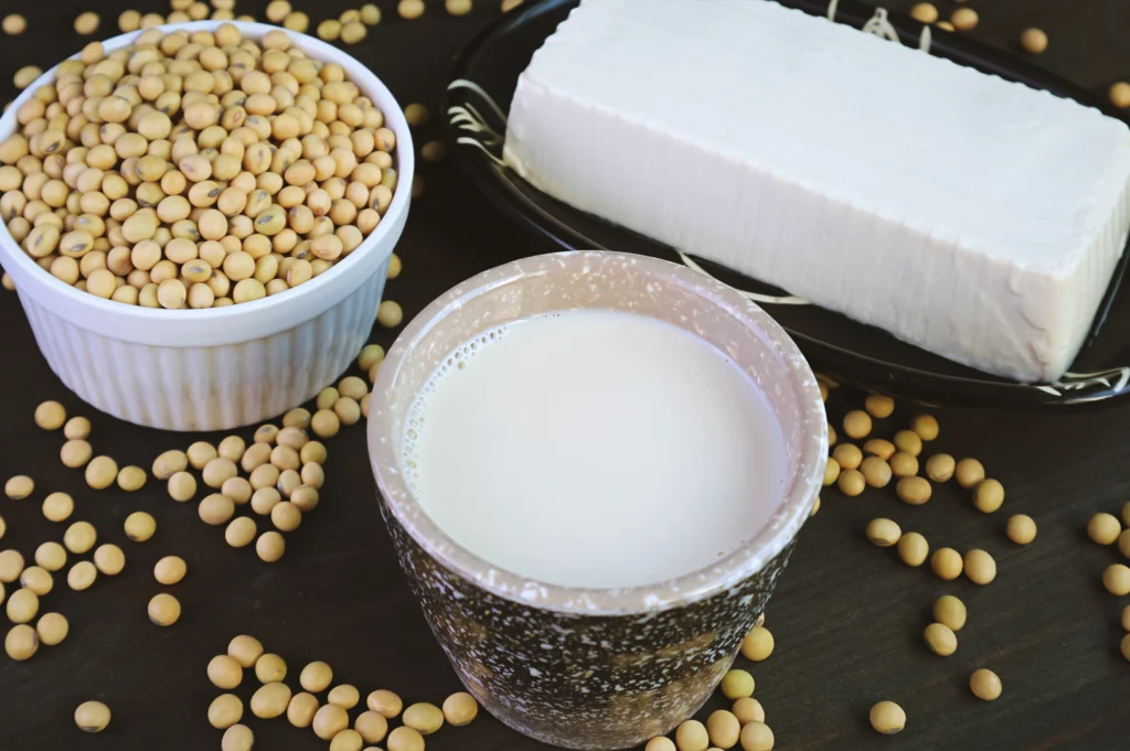 Eye-level close-up of soybeans, a glass of fresh soy milk, and a block of tofu arranged on a dark surface, illustrating light, protein-rich foods associated with balance and reduced richness fatigue.