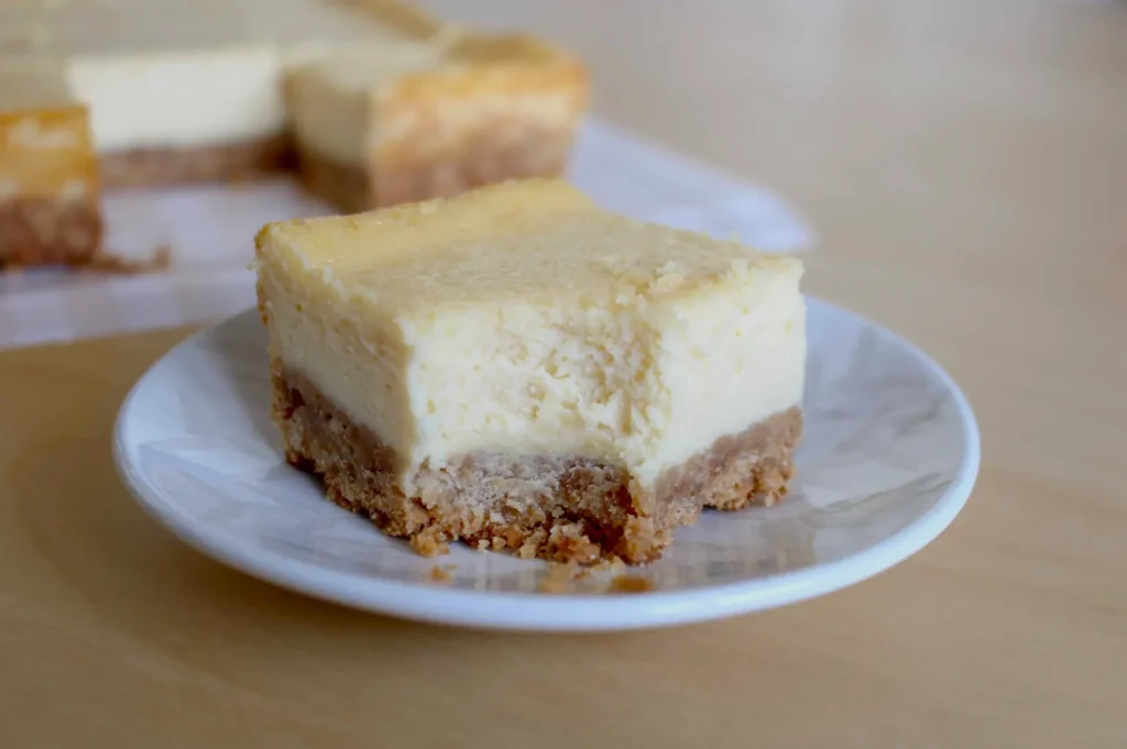 A slice of creamy cheesecake with a graham cracker crust sits on a white plate. It has a bite taken out, revealing a smooth texture. Another piece is in the background.