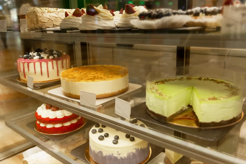 Straight-on eye-level display case shot of assorted whole cakes and cheesecakes arranged on glass shelves in a café dessert showcase, featuring multiple flavors and styles that reflect modern café culture.