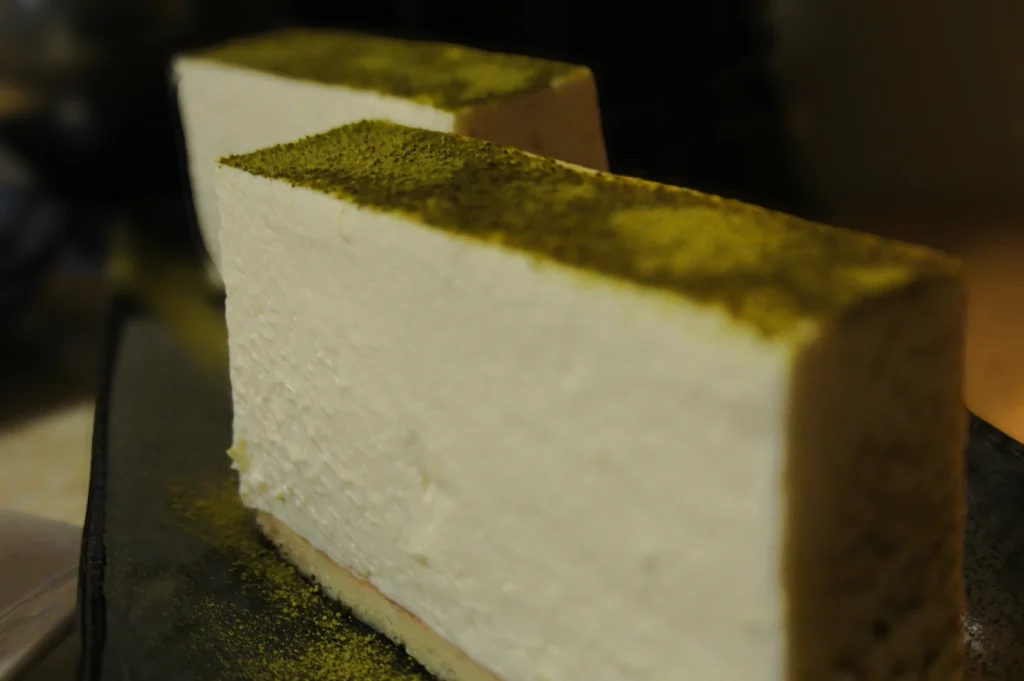 Close‑up side‑angle shot of sliced soy‑based cheesecake with a green topping, emphasizing creamy texture, clean layers, and minimalist dessert presentation.