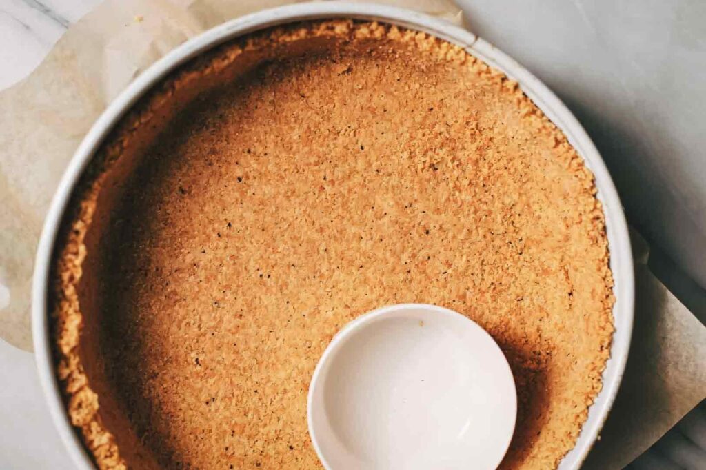 Overhead top-down shot of a baked crumb crust pressed evenly into a round cheesecake pan, showing a golden-brown texture before filling.