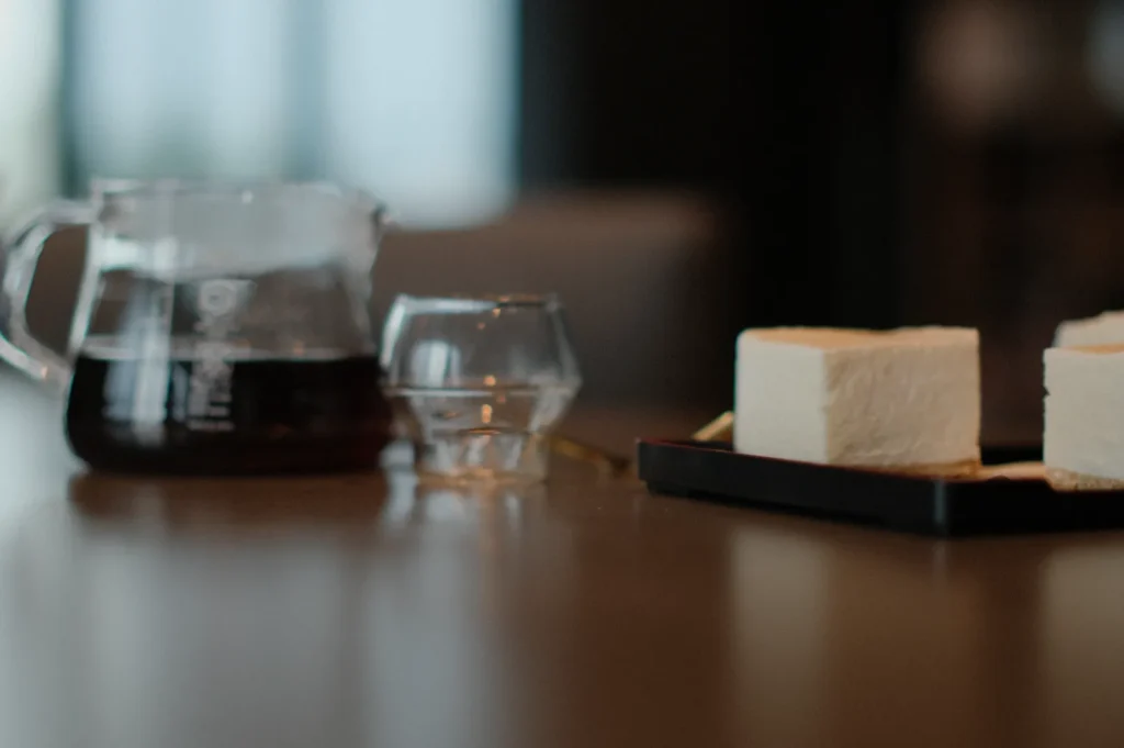 A glass pot of coffee sits beside a small clear glass on a wooden table. Two square slices of cake are placed nearby on a black tray, creating a cozy and inviting atmosphere.