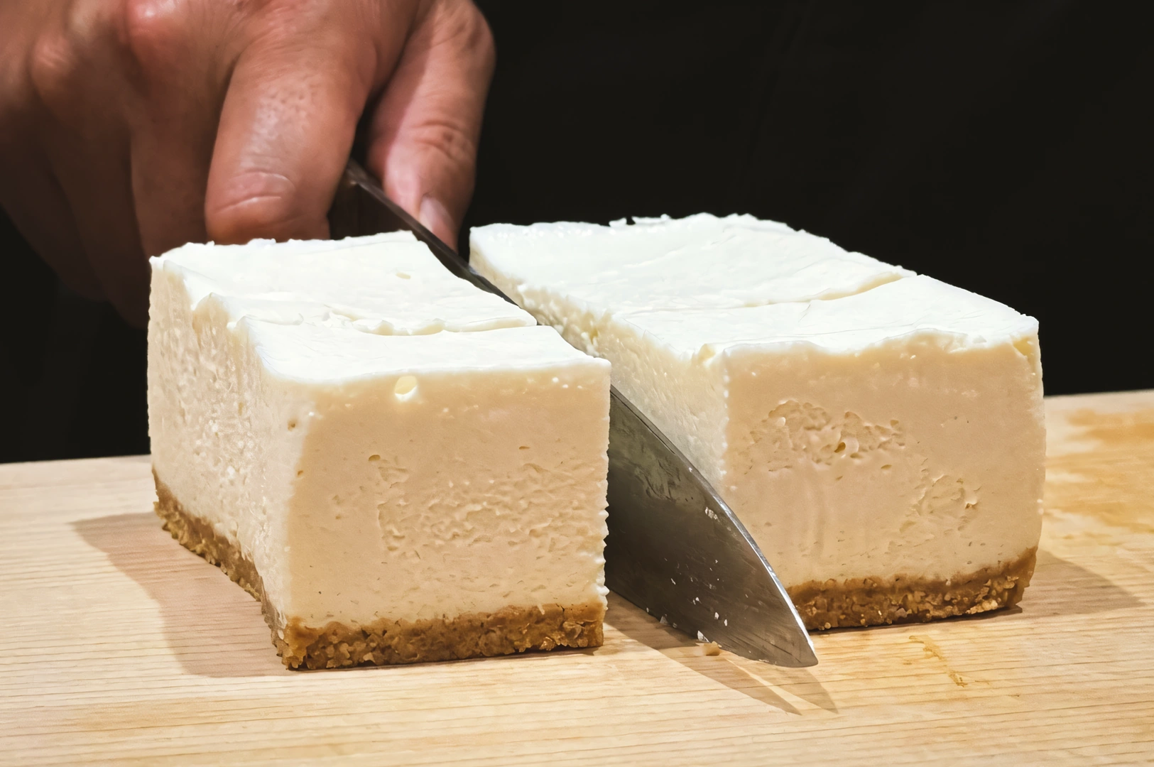 Close-up, eye-level shot of a knife slicing a creamy soy cheesecake with a smooth white filling and graham-style crust on a wooden cutting board.