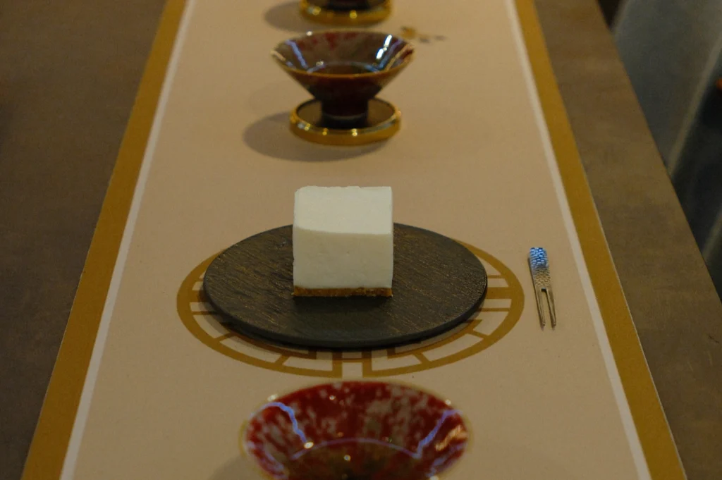 A slice of cheesecake on a dark plate is centered on an elegant table setting. Surrounding it are decorative red bowls and a small ornate fork. The scene conveys sophistication and anticipation.