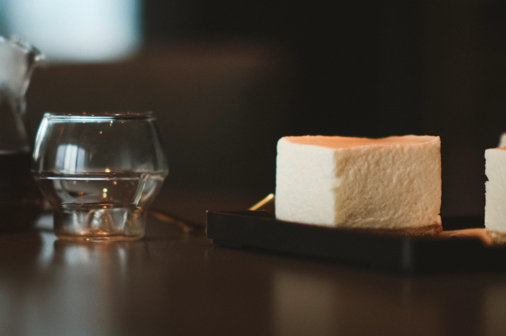 Eye-level close-up of a soy cheesecake dessert served on a dark tray beside a small glass cup, highlighting simplicity, restraint, and relief from overly rich flavors.