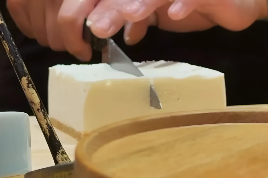 Hands carefully slice a block of tofu with a knife on a wooden cutting board, conveying focus and precision in a kitchen setting.
