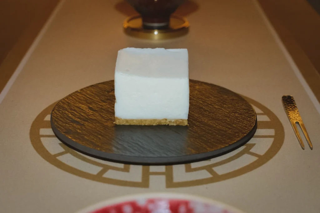 Straight-on eye-level shot of a square slice of soy cheesecake with a smooth white soy-based top and light golden crust, centered on a dark plate in a minimalist dessert presentation.