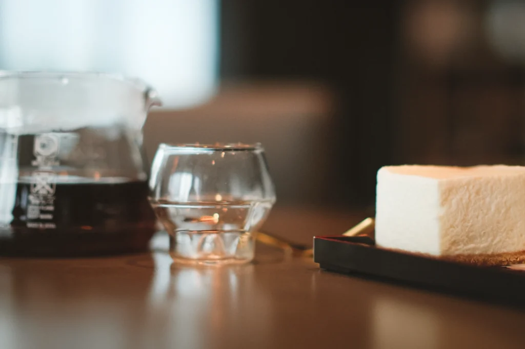 Low three-quarter close-up café table shot of a minimalist soy cheesecake block on a dark plate, paired with a glass of water and a coffee server in soft ambient lighting, highlighting dairy-free soy cheesecake texture, clean lines, and modern café dessert presentation.