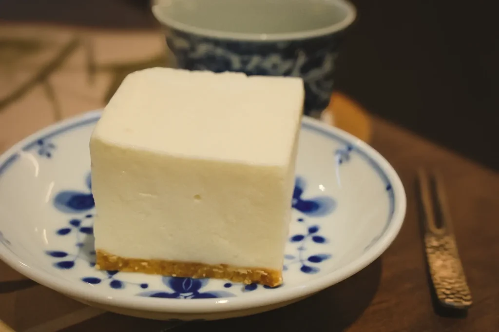 Eye-level three-quarter angle shot of a single square slice of soybean cheesecake on a ceramic plate with a minimal table setting in the background.