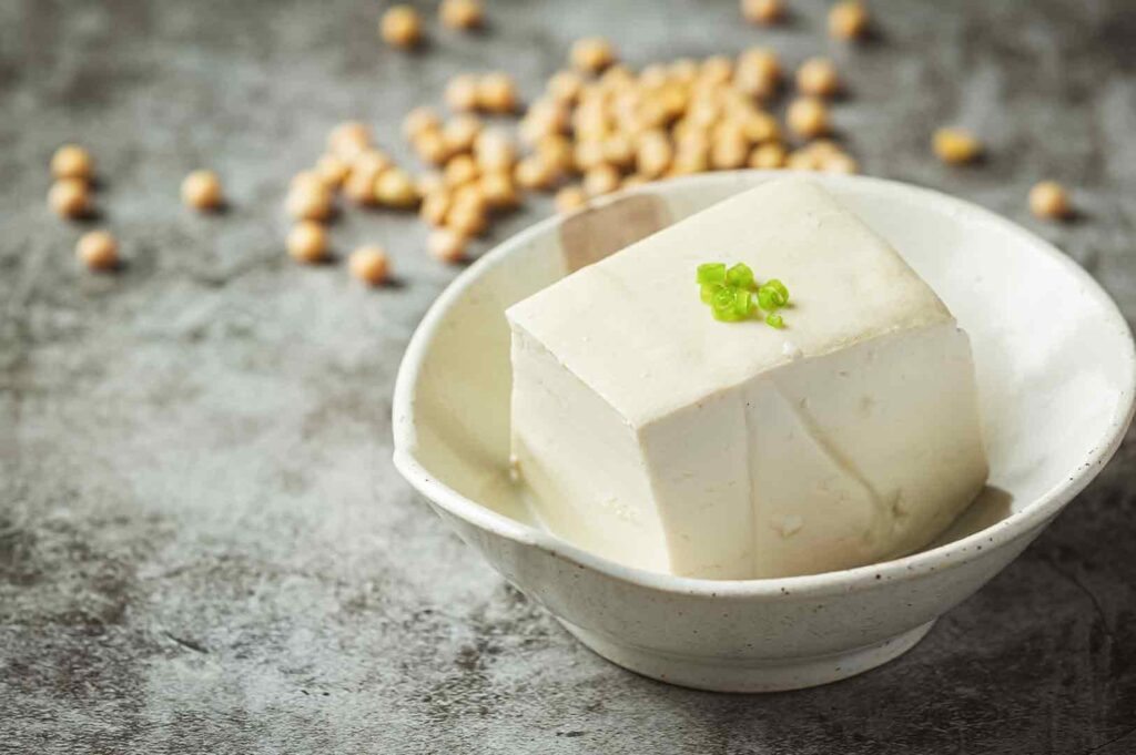 Three-quarter angle close-up shot of a block of silken tofu in a shallow bowl with soaking liquid, with whole soybeans scattered in the background on a gray surface.