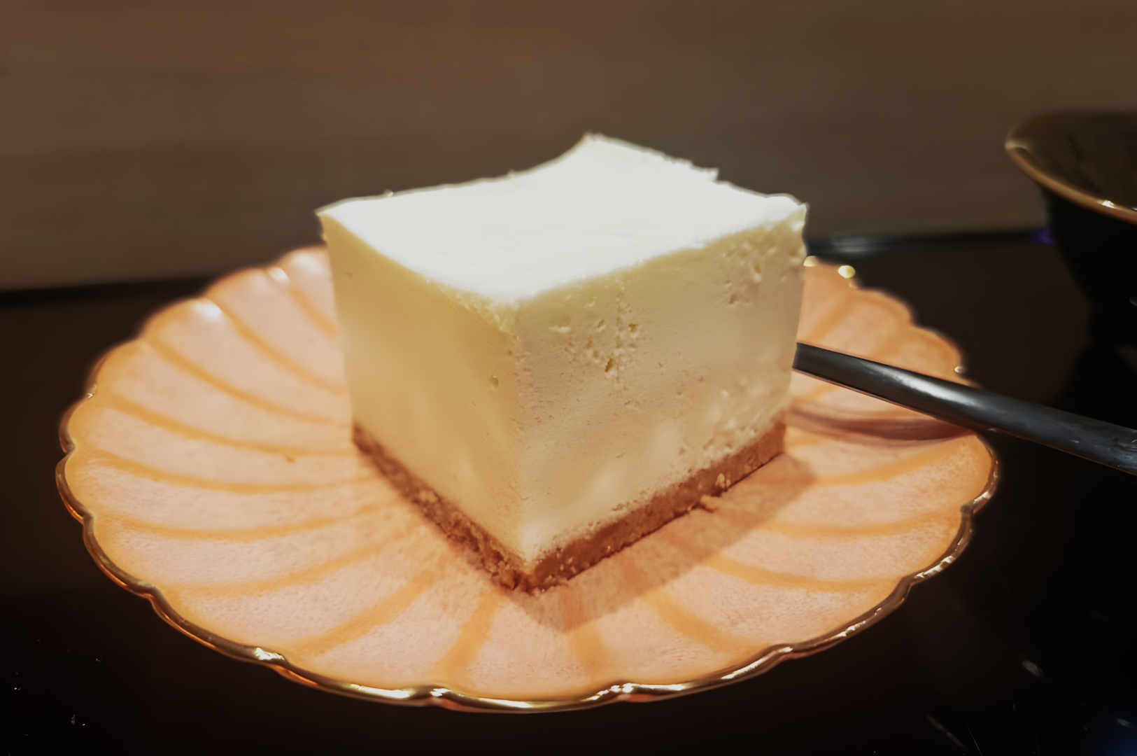 A slice of creamy cheesecake with a crumbly base on a pink, scalloped plate, accompanied by a fork. The mood is inviting and indulgent.