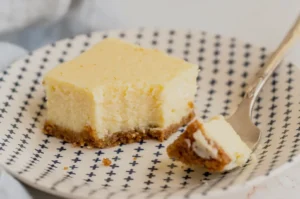 A slice of cheesecake with a graham cracker crust sits on a patterned plate. A fork beside it holds a bite-sized piece. The tone is inviting and delicious.
