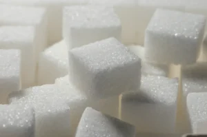 Close-up of a pile of white sugar cubes stacked unevenly, with fine grains visible. The image conveys a textured, orderly yet chaotic feel.