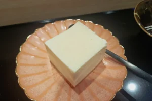 Square slice of white tofu on a round, pink scalloped plate, with a metallic spoon beside it. The setting is clean and minimal.