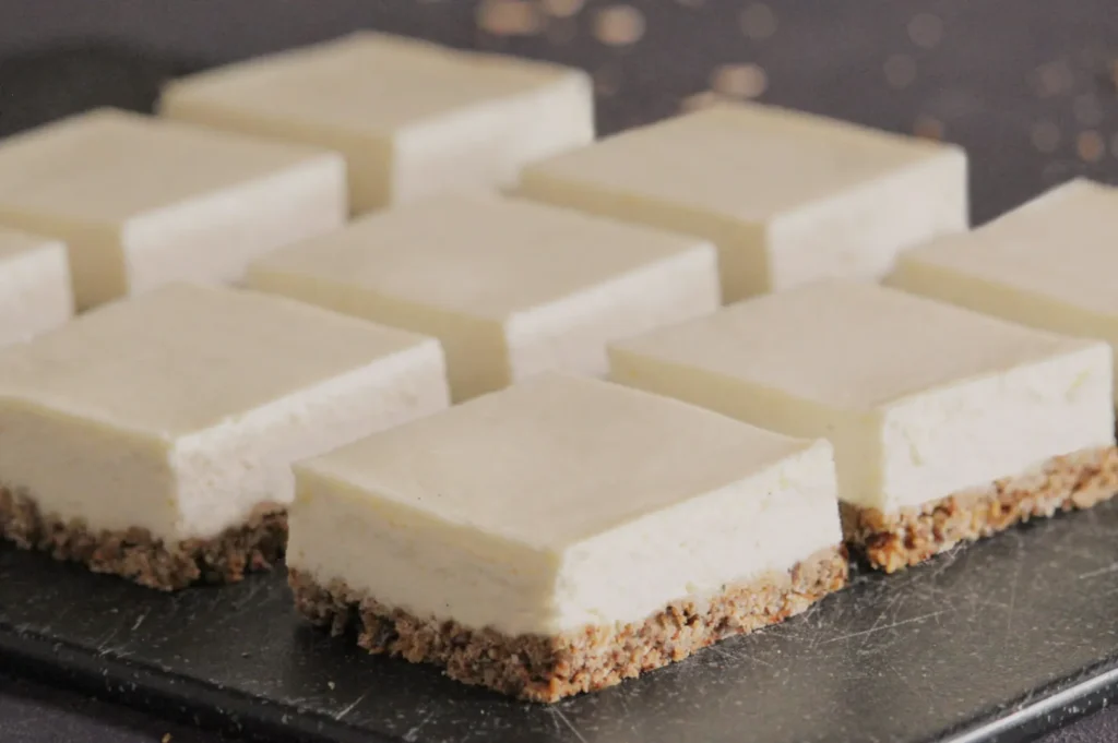 Nine square cheesecake bars with creamy tops and crumbly brown bases are neatly arranged on a dark slate, giving a fresh and inviting appearance.
