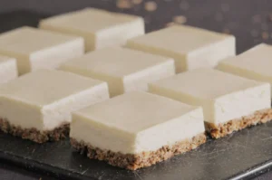 Nine square cheesecake bars with creamy tops and crumbly brown bases are neatly arranged on a dark slate, giving a fresh and inviting appearance.