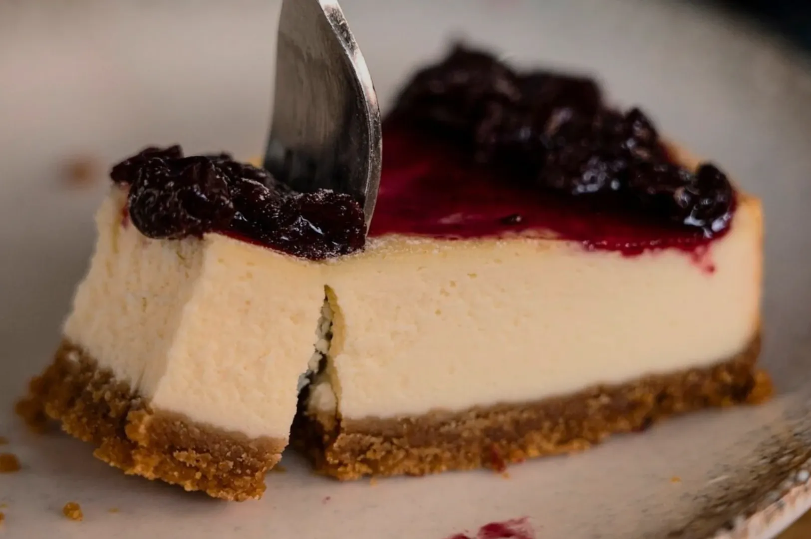 A slice of cheesecake with a golden-brown crust and creamy filling is topped with rich, glossy berry sauce. A fork cuts through the cake.