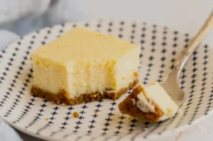 A creamy cheesecake square with a crumbly brown crust sits on a white plate with blue cross patterns. A fork holds a small cheesecake bite.