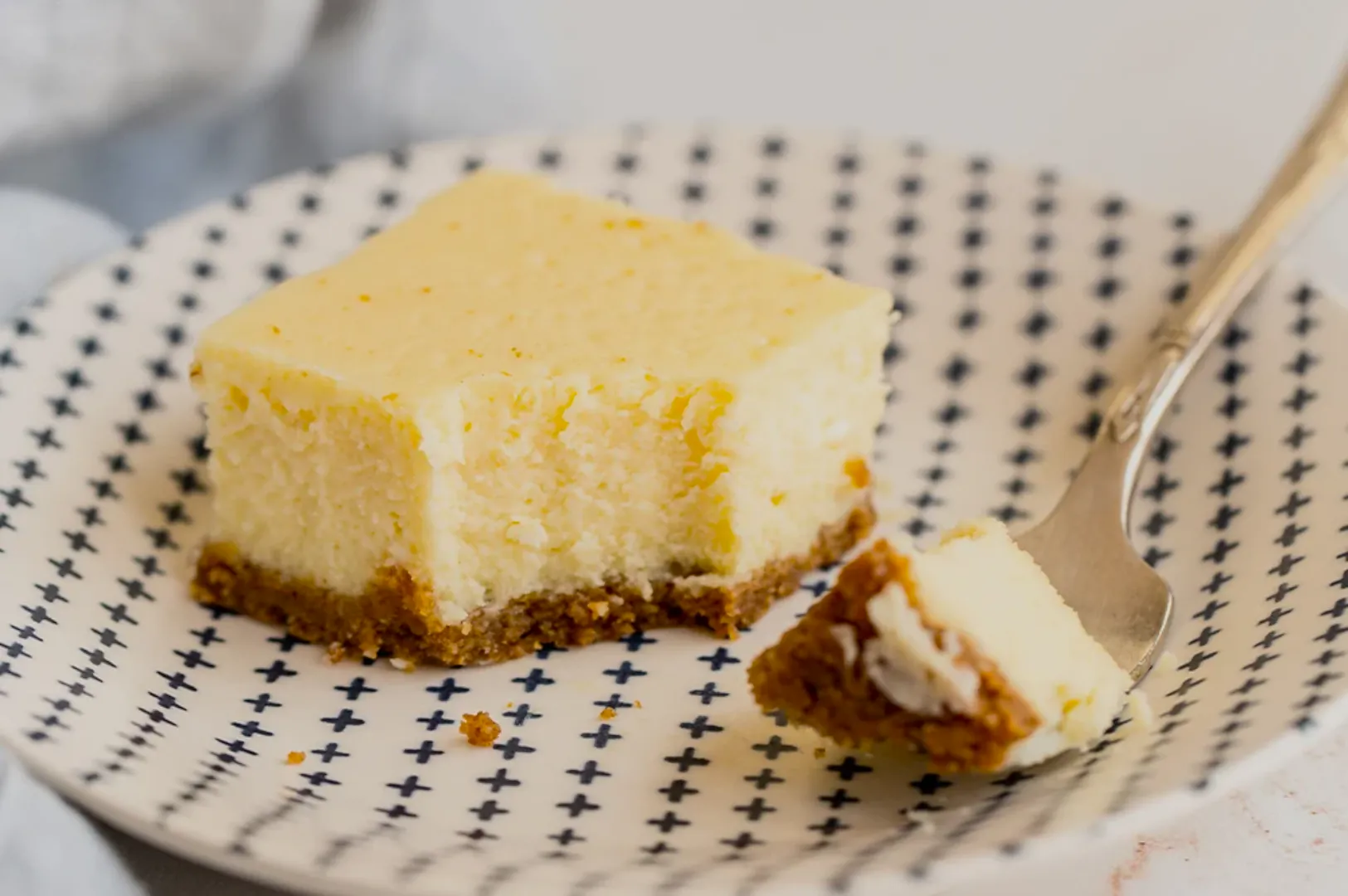 A creamy cheesecake square with a crumbly brown crust sits on a white plate with blue cross patterns. A fork holds a small cheesecake bite.
