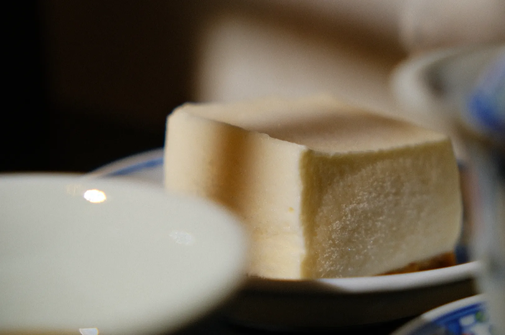 A close-up of a butter cube on a small dish, partially obscured by out-of-focus white plates. The lighting is soft, creating a warm, inviting tone.