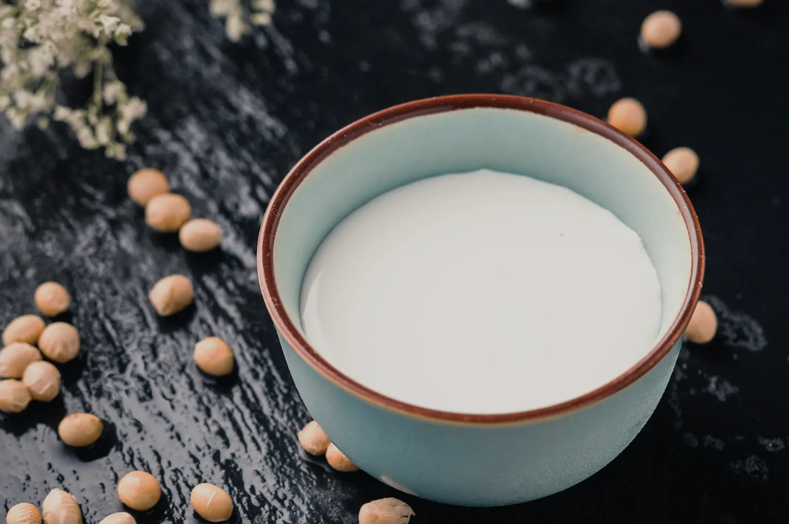 A blue ceramic bowl filled with soy milk sits on a black textured surface, surrounded by scattered soybeans and delicate white flowers, creating a serene, natural scene.Close-up of numerous beige soybeans scattered on a textured, off-white surface. The composition appears natural and earthy.