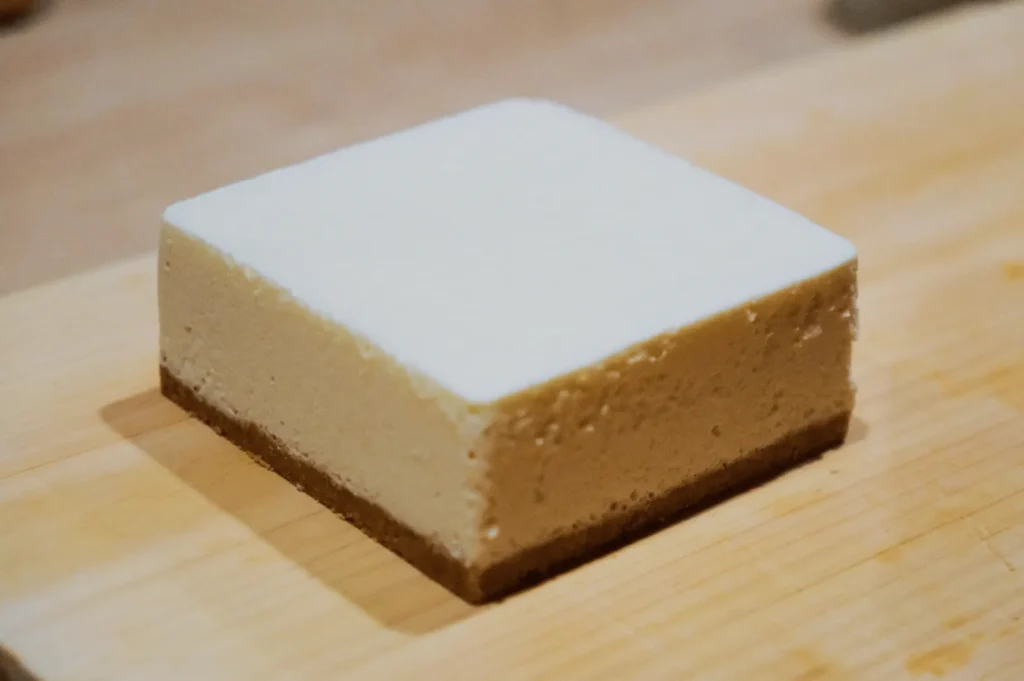 A square slice of cheesecake sits on a wooden board. It has a creamy, smooth top layer over a firm, slightly crumbly biscuit base, evoking a comforting, indulgent mood.