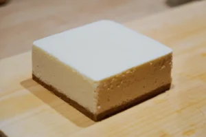 A square slice of cheesecake sits on a wooden board. It has a creamy, smooth top layer over a firm, slightly crumbly biscuit base, evoking a comforting, indulgent mood.