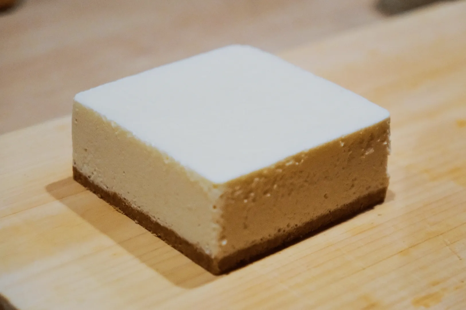 A square slice of cheesecake sits on a wooden board. It has a creamy, smooth top layer over a firm, slightly crumbly biscuit base, evoking a comforting, indulgent mood.